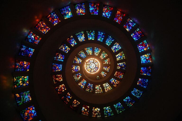 A low-angle view looking up at a dark spiral ceiling where a central bright light illuminates a sequence of colorful stained-glass windows, representing a scalar value bringing light to an abstract space.