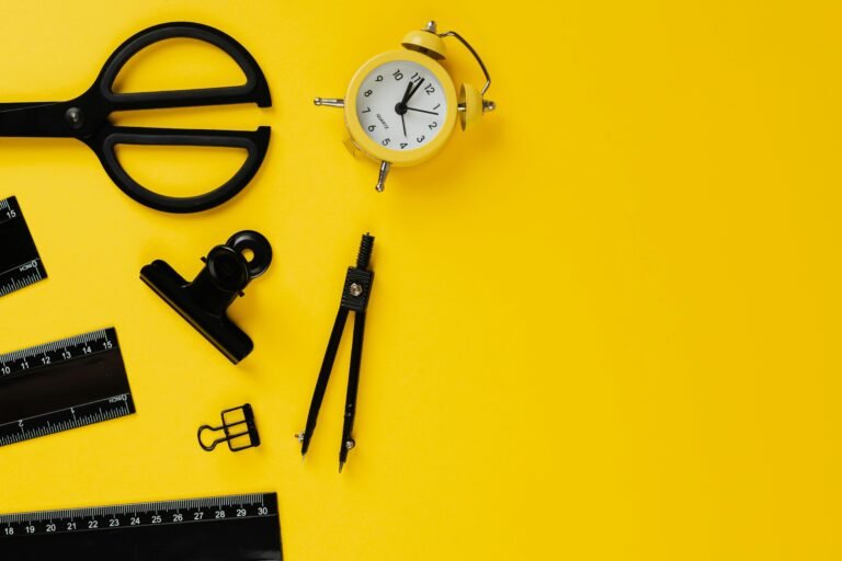 A top-down view of mathematical and drafting tools—including a compass, rulers, scissors, and a yellow alarm clock—laid out on a vibrant yellow background. (Alt text should be descriptive for accessibility and screen readers.)